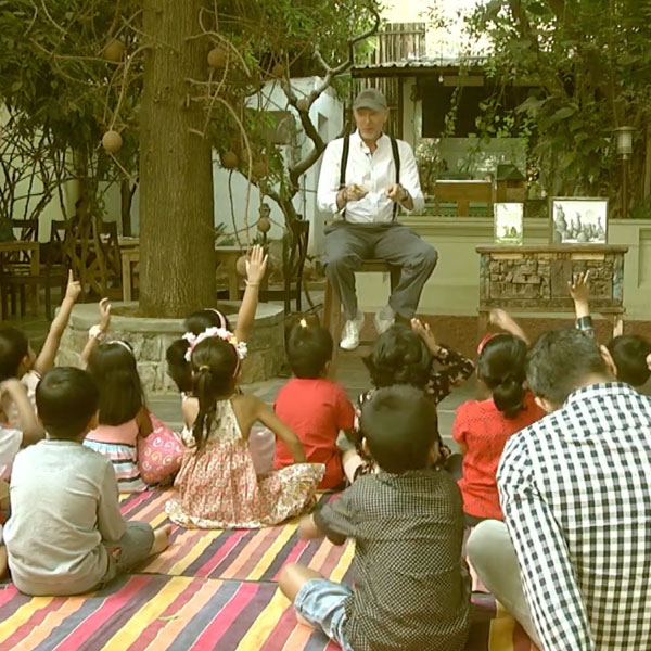 man sitting on a stool, reading to a group of kids, and a parent, sitting on a blanket watching him