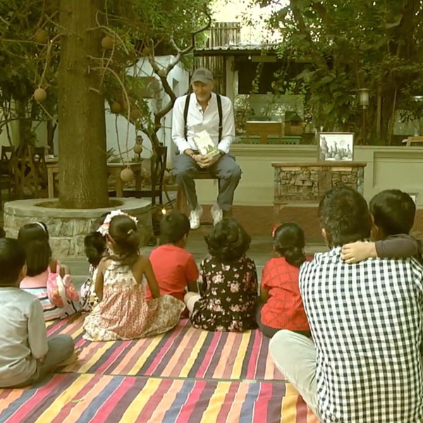 man sitting on a stool, reading to a group of kids, and a parent, sitting on a blanket watching him
