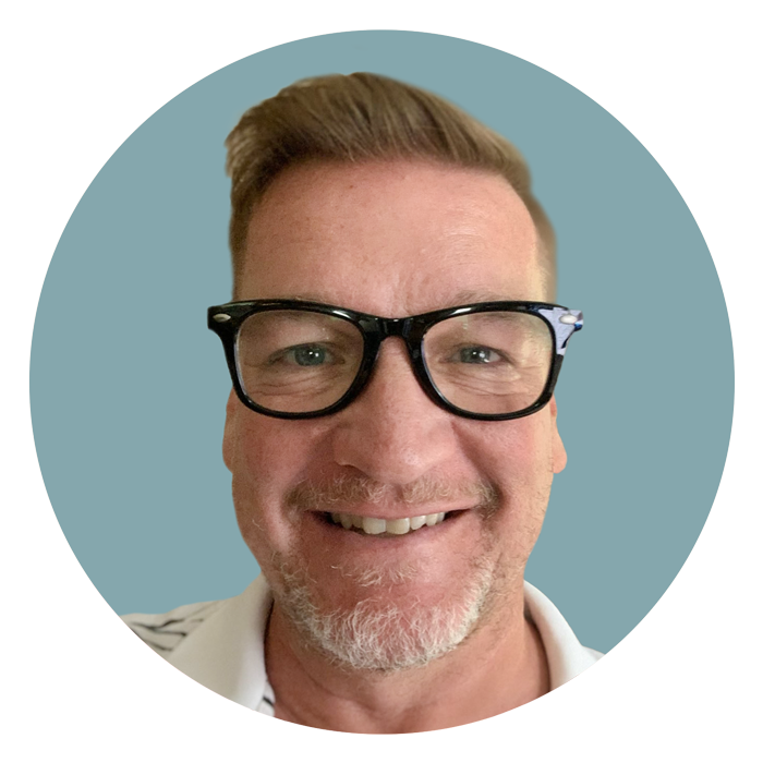 Man with blonde low fade hair wearing black-framed glasses smiling